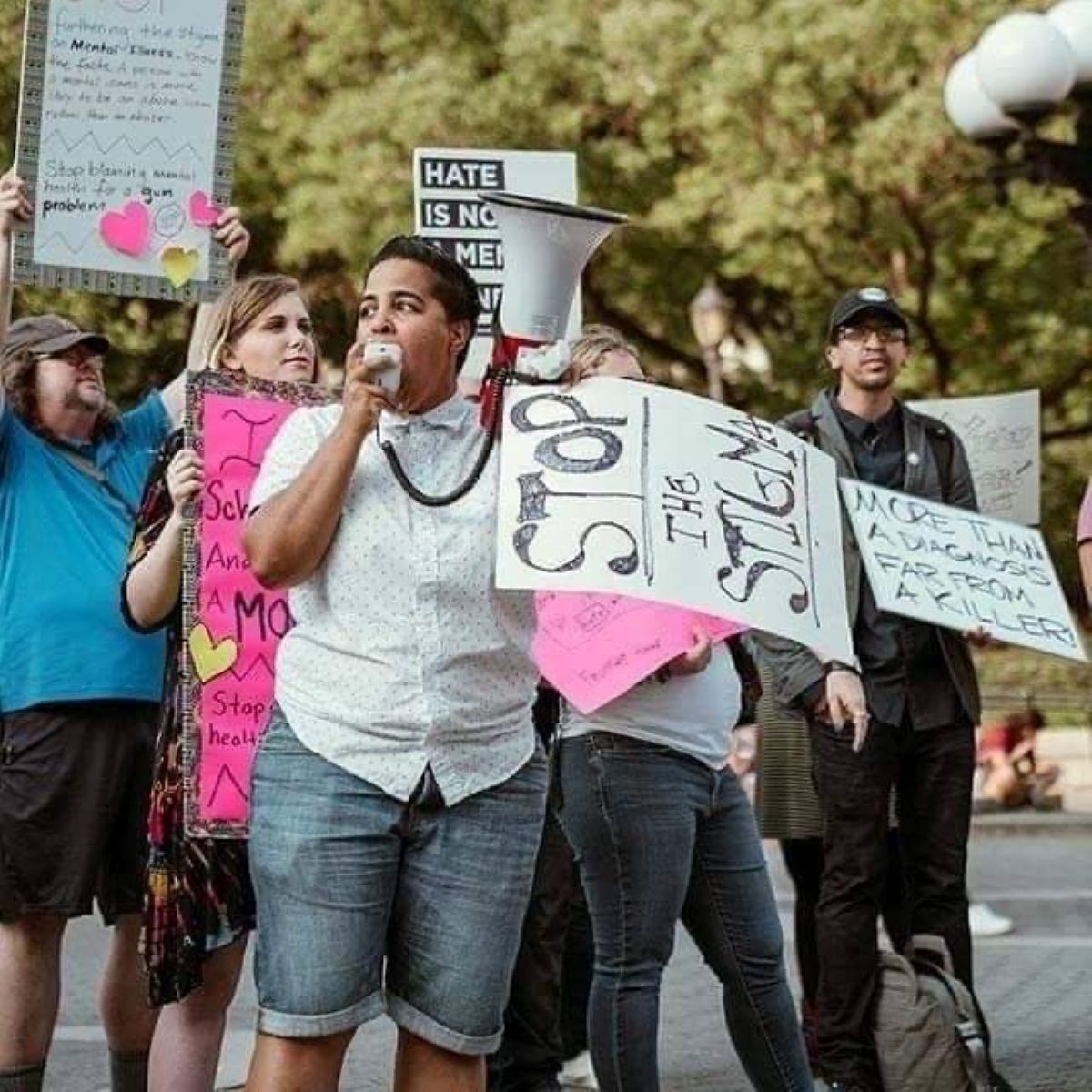 A brown, queer, non-binary person, outdoors, holds a megaphone. They are wearing a dressy white shirt and denim shorts. They have short black, spiky hair. Four people behind them hold signs, one reads Stop the stigma.