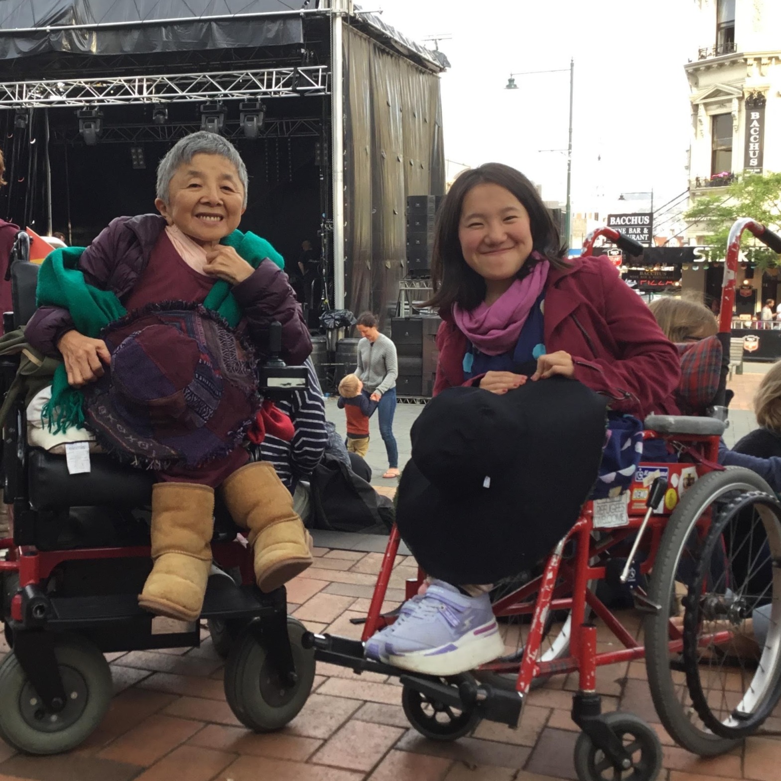 A Japanese mother and daughter both using wheelchairs. They sit in front of a New Year's Concert stage in the city centre.