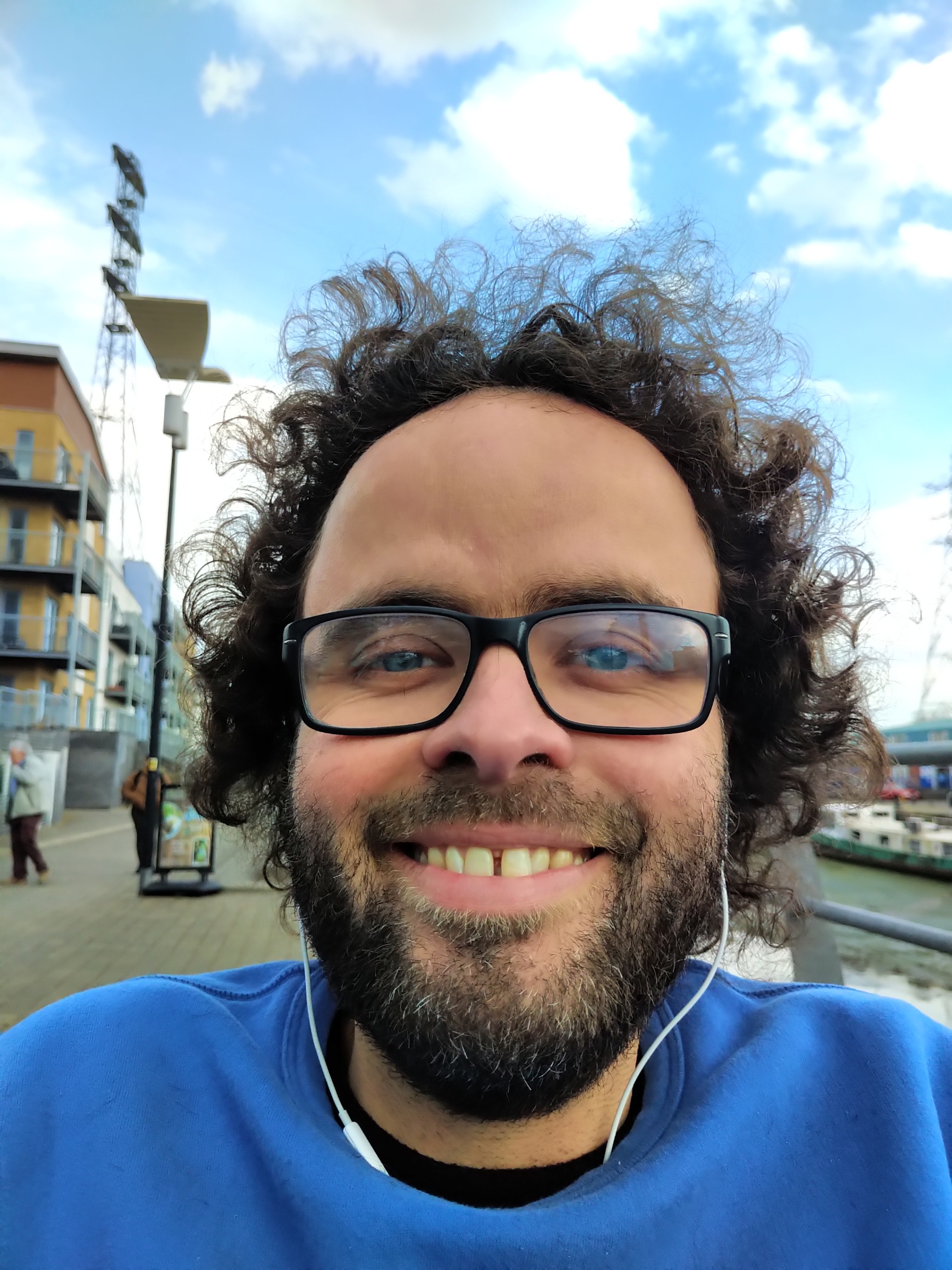 Peter smiles, a white man with dark curly hair frazzling skywards, beard with specks of grey, black glasses. He’s wearing a dark blue jumper and the sky is a light blue with a touch of cloud behind him”.